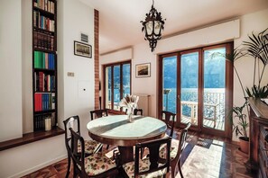 A dining area with a round table, chairs, a bookshelf, and a view of the outdoors through large windows. - Villa Fisogni - La Sonnambula (Moltrasio)