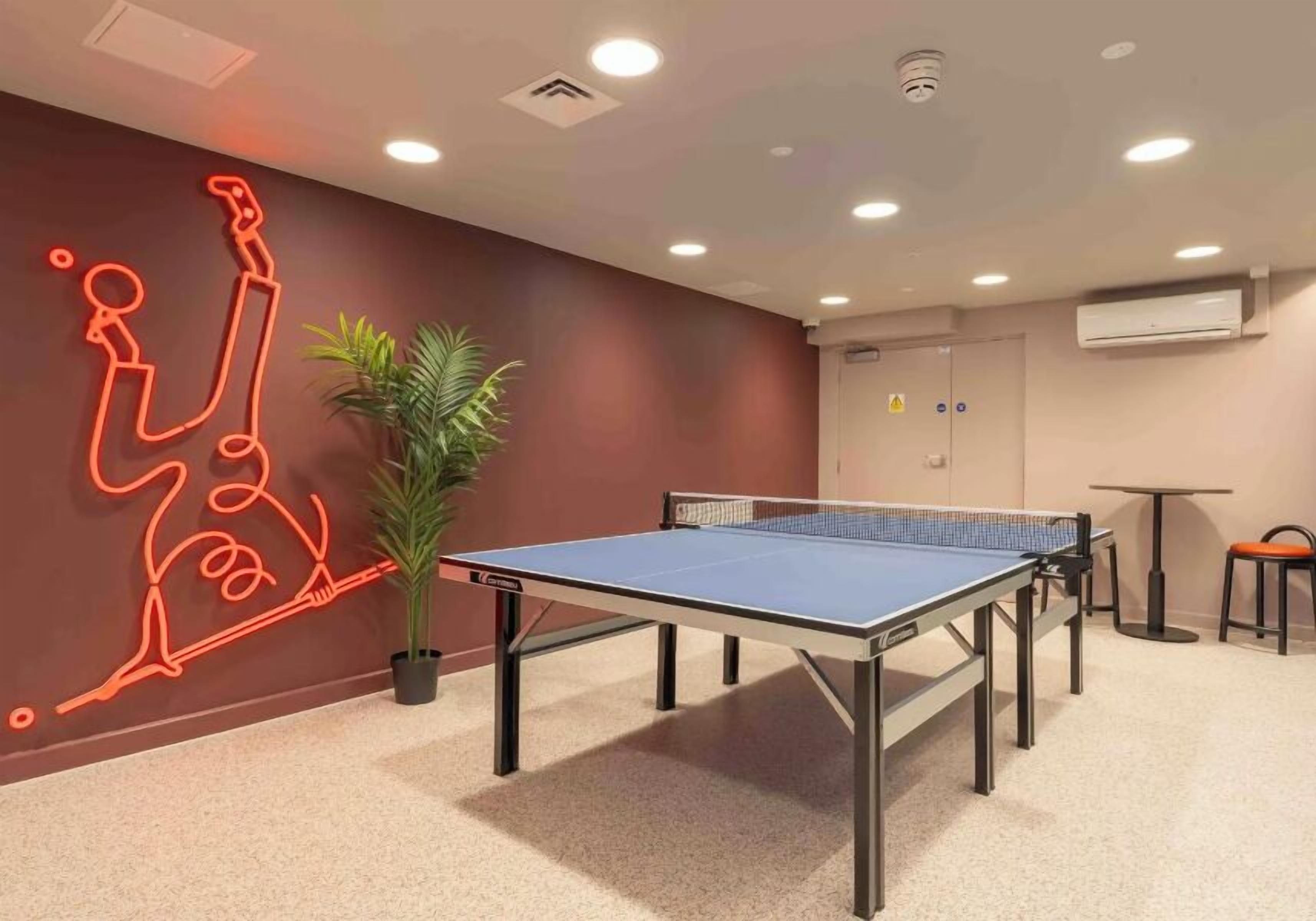 A room with a ping pong table, a neon art installation on the wall, a potted plant, and a door with a safety sign.