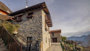 Edifici de pedra amb una escala que condueix a una porta de fusta, envoltat d'un camí de pedra i vistes a les muntanyes.