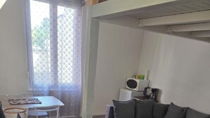 Ein Wohnzimmer mit Sofa, Glastisch, Fernseher, Esstisch mit Stühlen und einem fensterbepflanzten Fenster.