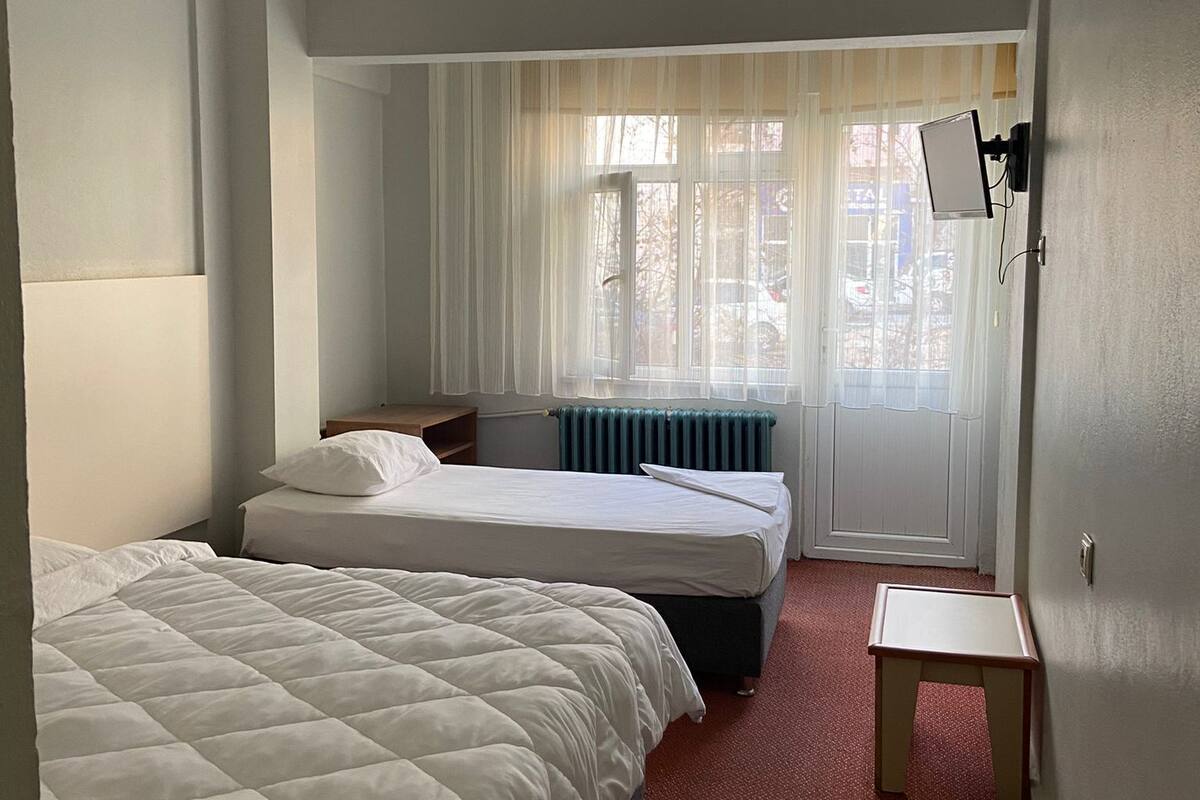 A hotel room with two beds, a small table, a radiator, and a window with sheer curtains.