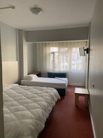 Quarto de hotel com duas camas, uma mesinha, um radiador e uma janela com cortinas leves.