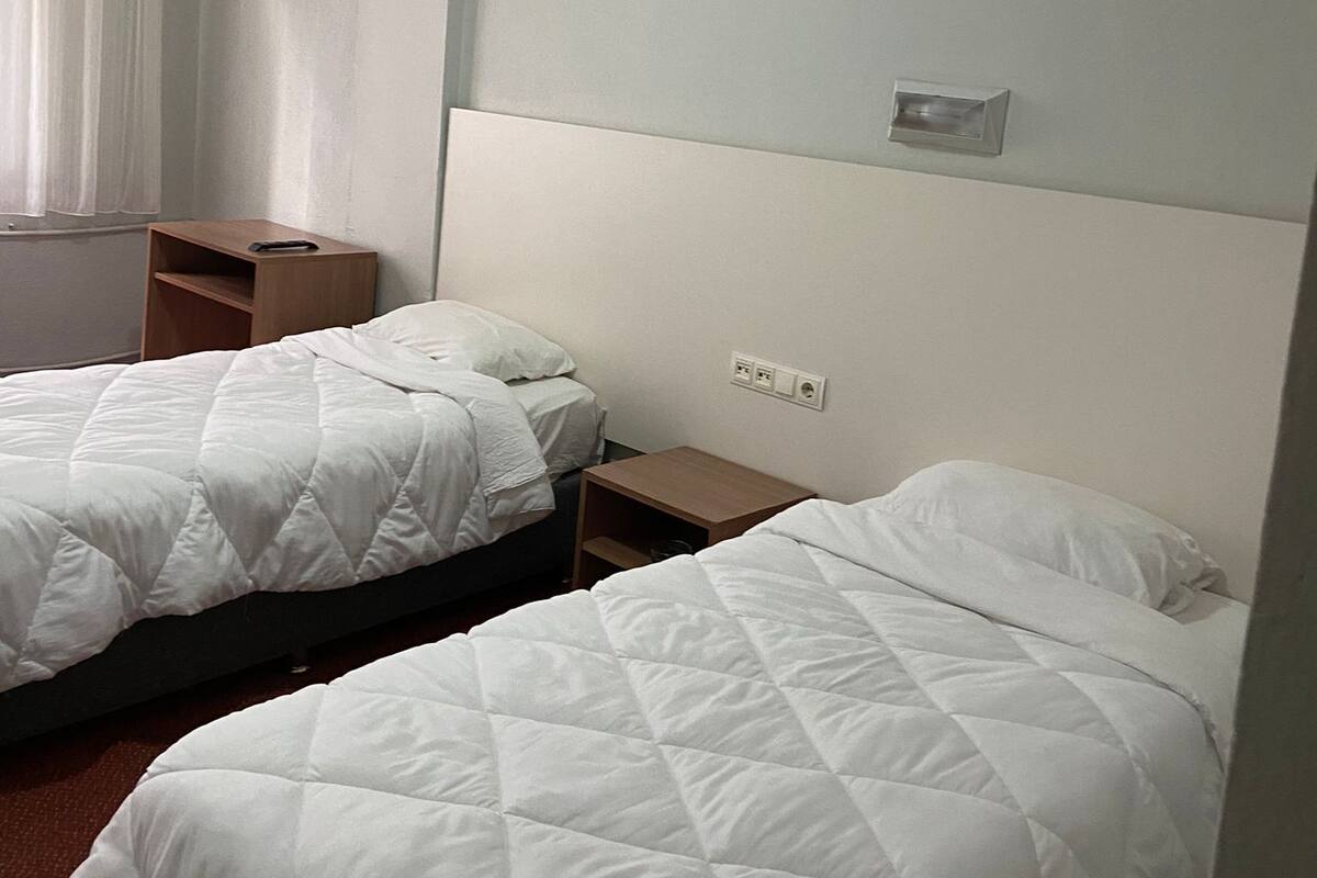 A hotel room with two beds, a nightstand, a wall socket, and a window with curtains.