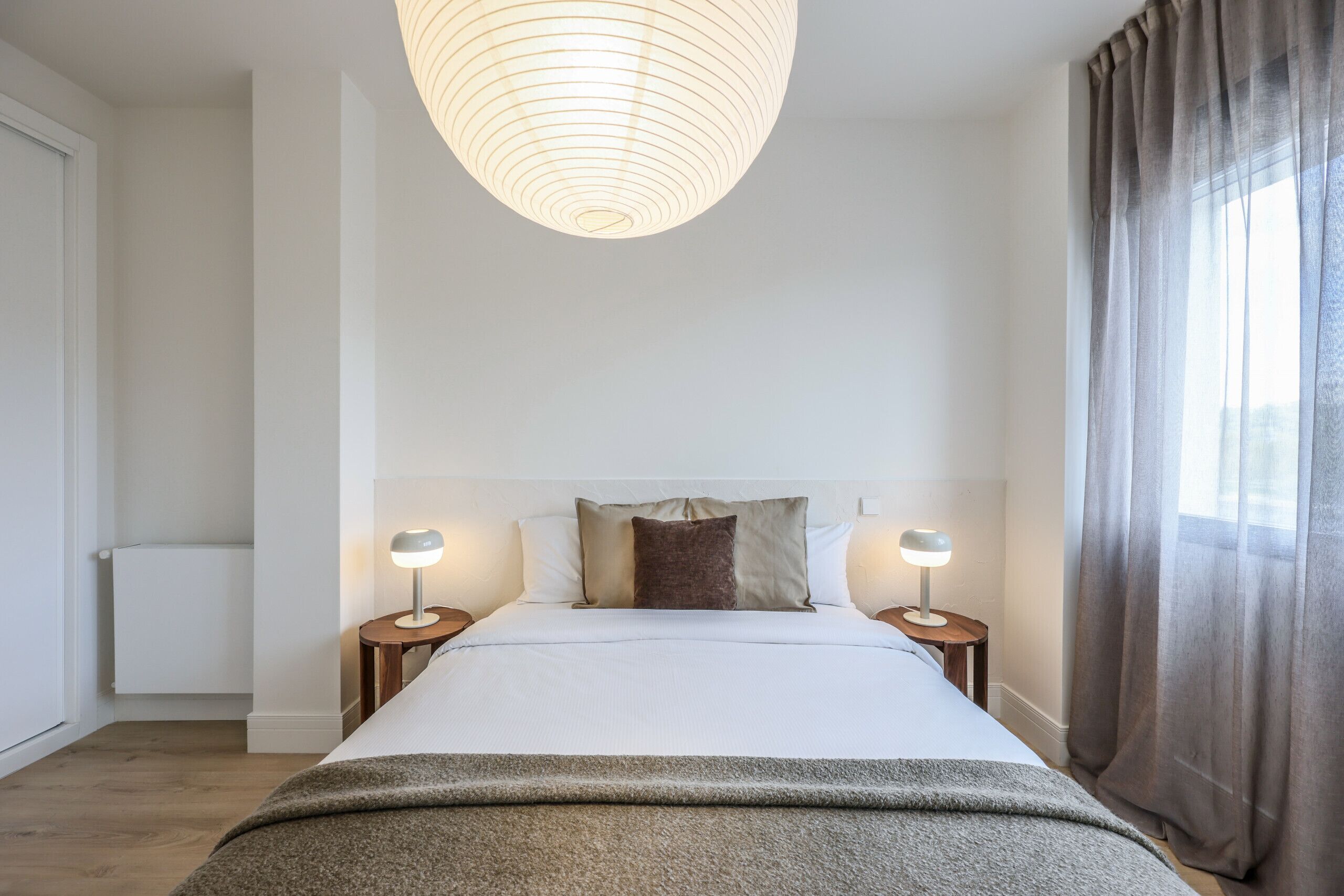 A modern hotel room with a large bed, two bedside tables with lamps, a large window with curtains, and a unique ceiling light fixture.