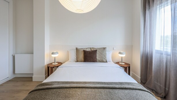 Una habitación de hotel moderna con una cama grande, dos mesitas de noche con lámparas, un ventanal con cortinas y una lámpara de techo original.