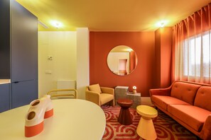 Una habitación con una mesa redonda, un sofá, una silla, un taburete pequeño, un espejo y una ventana con cortinas.