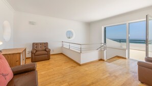 A spacious room with a wooden floor, a brown leather chair, a small wooden table, and a large sliding glass door leading to a balcony with a view of the sea. - Holiday Home 'Ocean Villa Ericeira' with Sea View, Private Terrace and Wi-Fi (Ericeira)