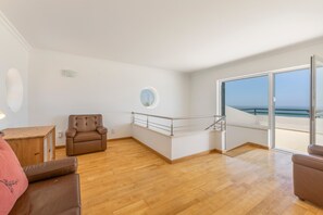 A spacious room with a wooden floor, a brown leather chair, a small wooden table, and a large sliding glass door leading to a balcony with a view of the sea. - Holiday Home 'Ocean Villa Ericeira' with Sea View, Private Terrace and Wi-Fi (Ericeira)