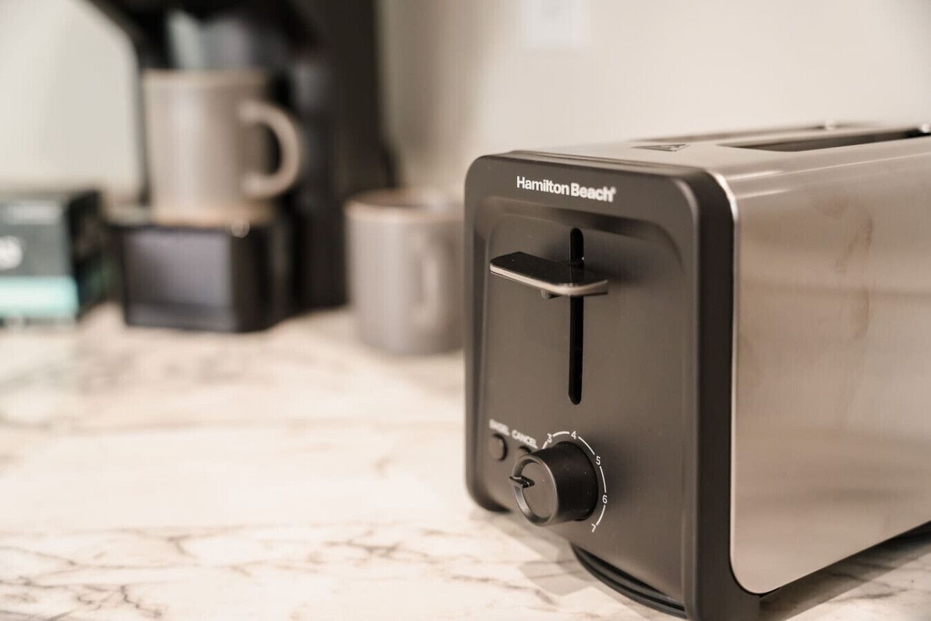 A Hamilton Beach toaster on a marble countertop with a coffee maker and mug in the background.