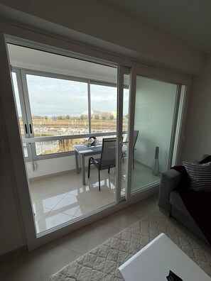 Ein modernes Wohnzimmer mit einem großen Schiebeglasfenster, das auf einen Balkon führt. Das Zimmer verfügt über ein graues Sofa, einen weißen Couchtisch und einen Esstisch mit Stühlen.