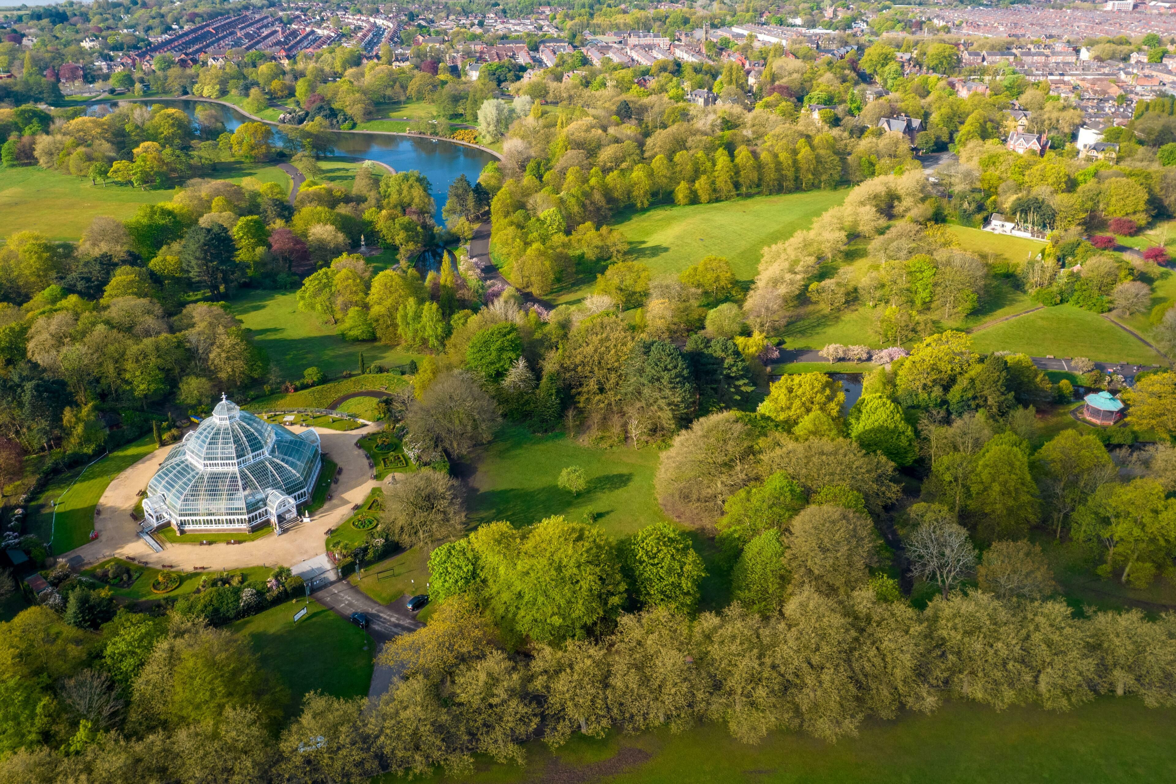 Luchtbeeld van een park met een groot glazen conservatorium, omringd door weelderige begroeiing en een meer.