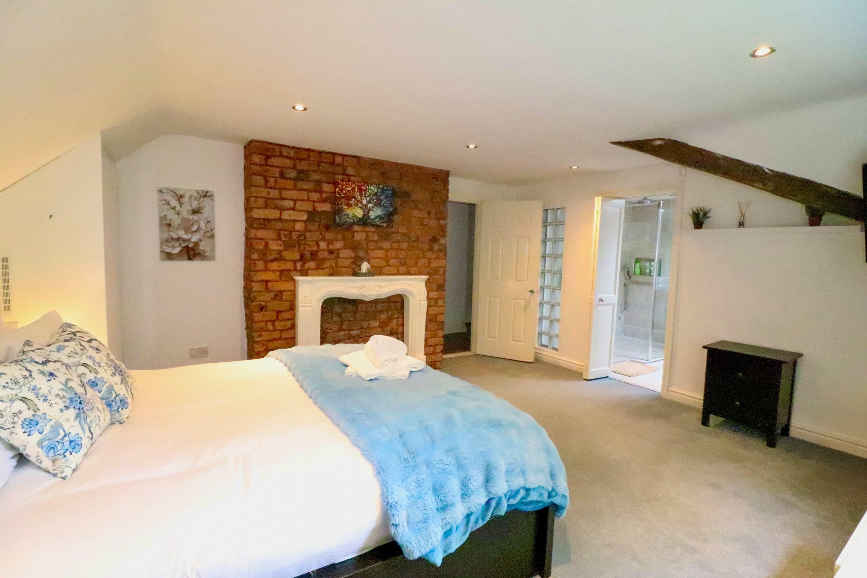 A bedroom with a large bed, a fireplace, a door leading to a bathroom, and a small table.