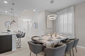 A modern kitchen with a central island, a dining area with a white table set for six, and a staircase leading to another level. - 7BR Home for 15 w/ Private Pool – Windsor Cay (Clermont)