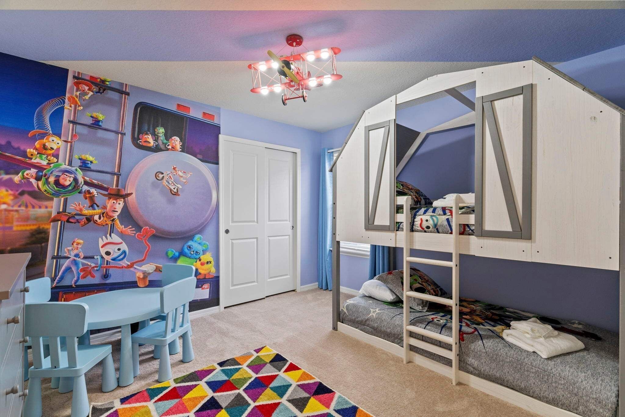 Habitación infantil con literas, una alfombra colorida, un escritorio con sillas y un mural en la pared.
