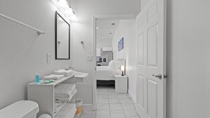 Et moderne badeværelse med toilet, håndvask og håndklæderadiator. Et spejl er monteret over håndvasken, og der er en lampe over spejlet. Badeværelset giver adgang til et soveværelse med en seng og et natbord med en lampe.
