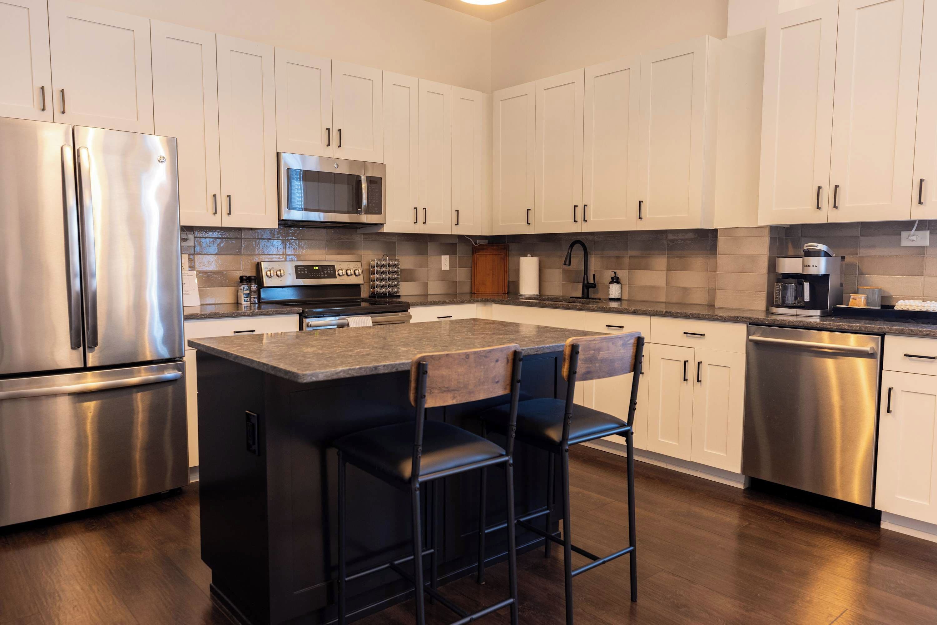 A modern kitchen with stainless steel appliances, a central island with bar stools, and a tiled backsplash.