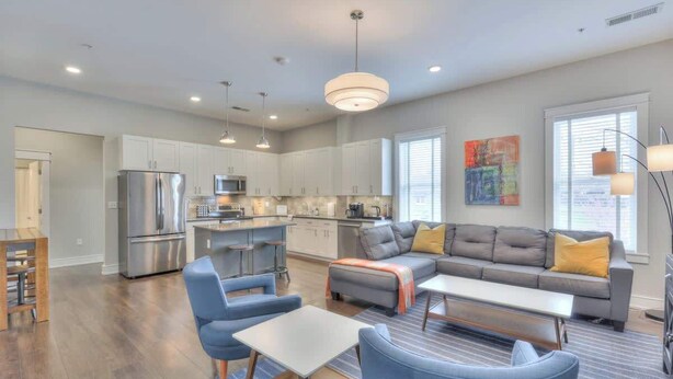 A modern kitchen with stainless steel appliances, a dining area with blue chairs and a white table, and a living room with a gray sofa and yellow pillows.