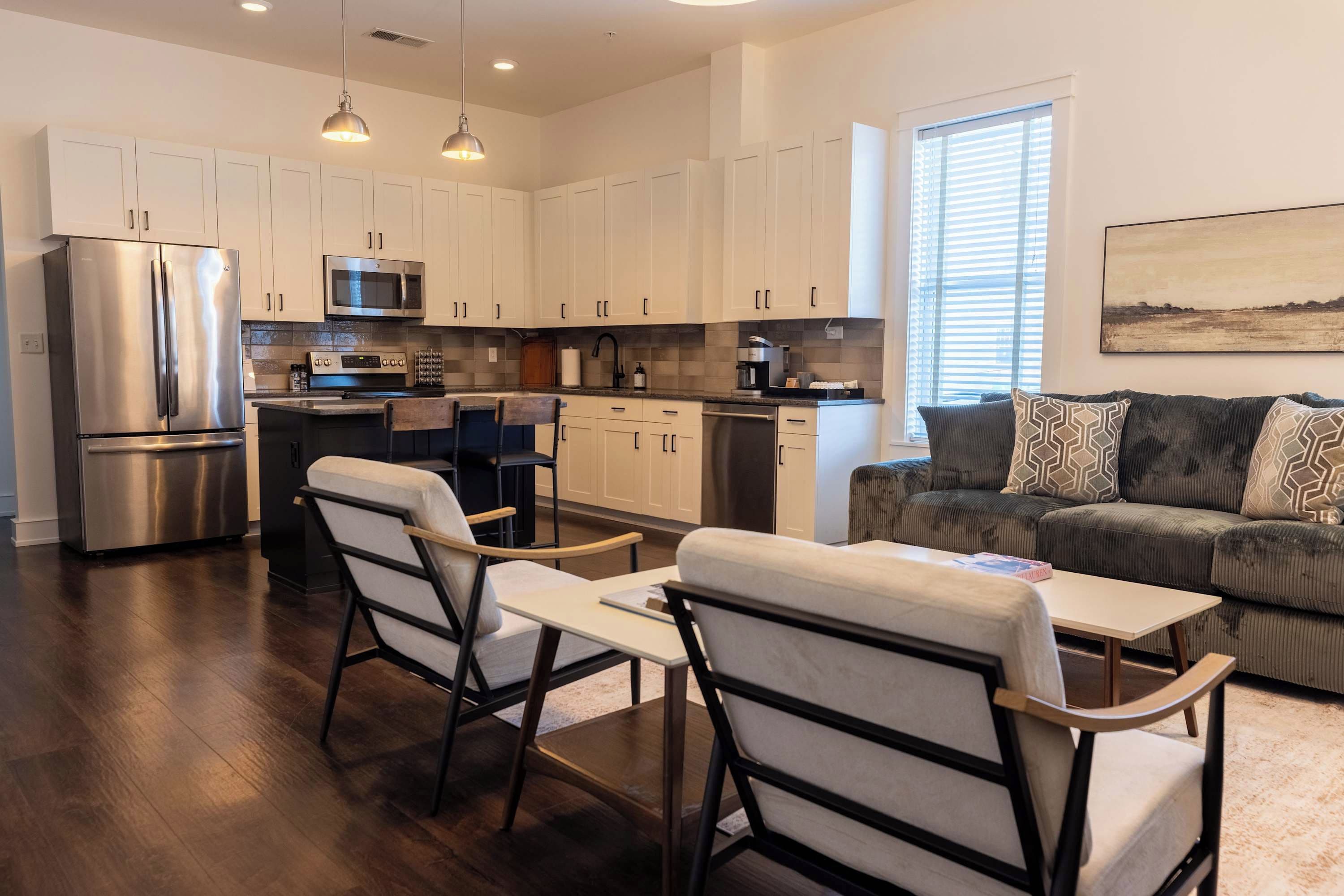 A modern kitchen with stainless steel appliances, a dining area with a table and chairs, and a living room with a sofa and a painting.