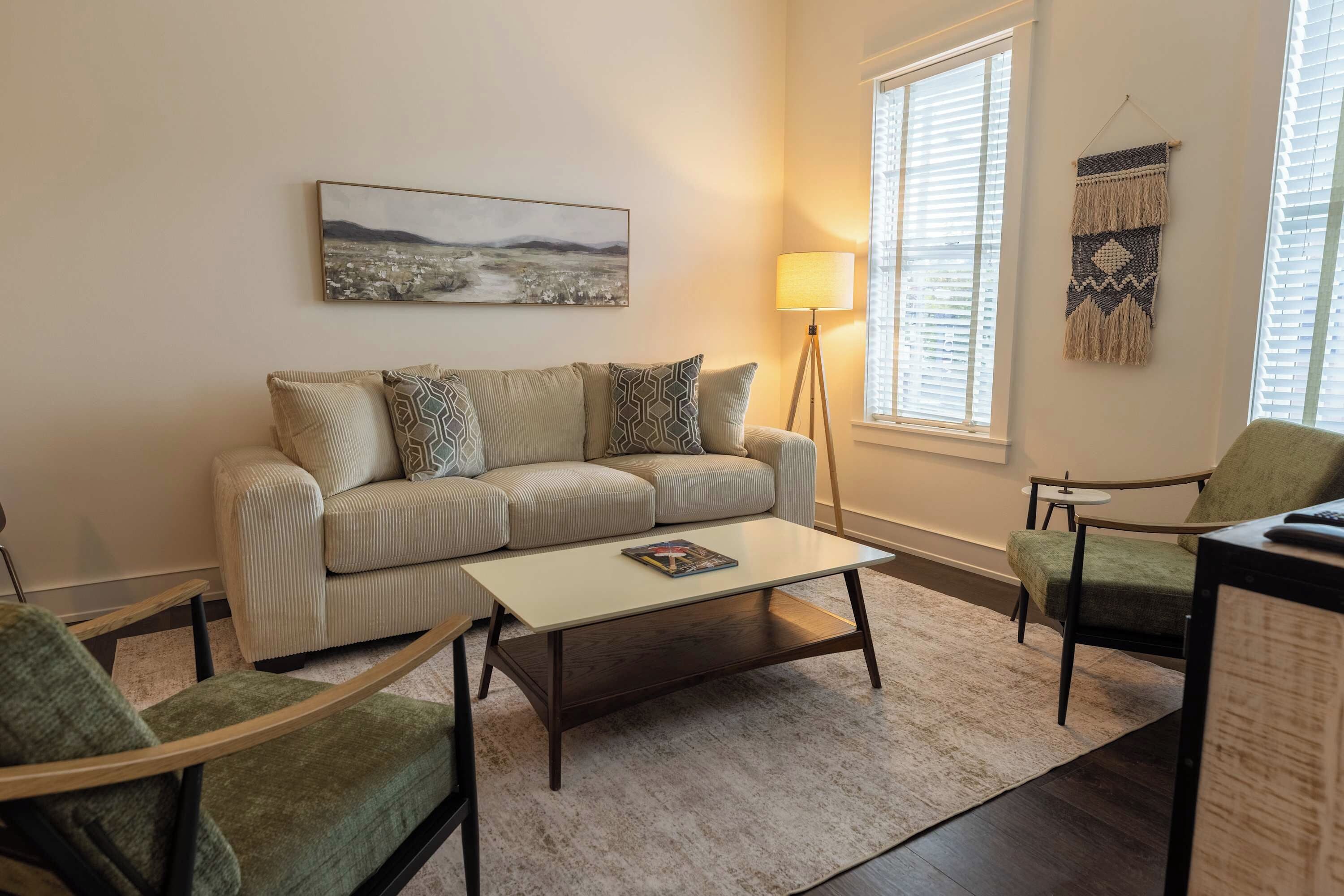 Een woonkamer met een bank, twee fauteuils, een salontafel, een vloerlamp, een schilderij en een raam met jaloezieën.