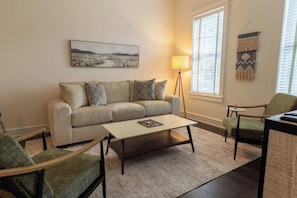 A living room with a sofa, two armchairs, a coffee table, a floor lamp, a painting, and a window with blinds. - 1-BR Condo in 12 South I Unit 202 (Nashville)