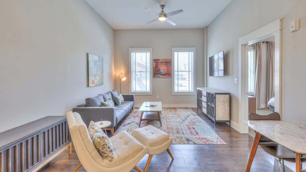 A spacious living room with a sofa, ottomans, a coffee table, and a dining table. There are large windows, a ceiling fan, and artwork on the walls. - 1-BR Condo in 12 South I Unit 202 (Nashville)