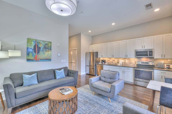 A modern living room with a grey sofa, armchair, and coffee table. The kitchen features white cabinets, stainless steel appliances, and a dining area with a table and chairs.