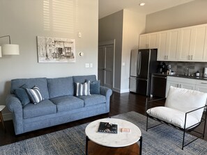 Ein Wohnzimmer mit Sofa, Sessel und Couchtisch, eine Küche mit Geräten und eine Tür zu einem weiteren Raum.