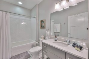 A modern bathroom with a white bathtub, a large mirror, and a vanity with a sink and toiletries. - 1-BR Studio Condo in 12 South I Unit 203 (Nashville)