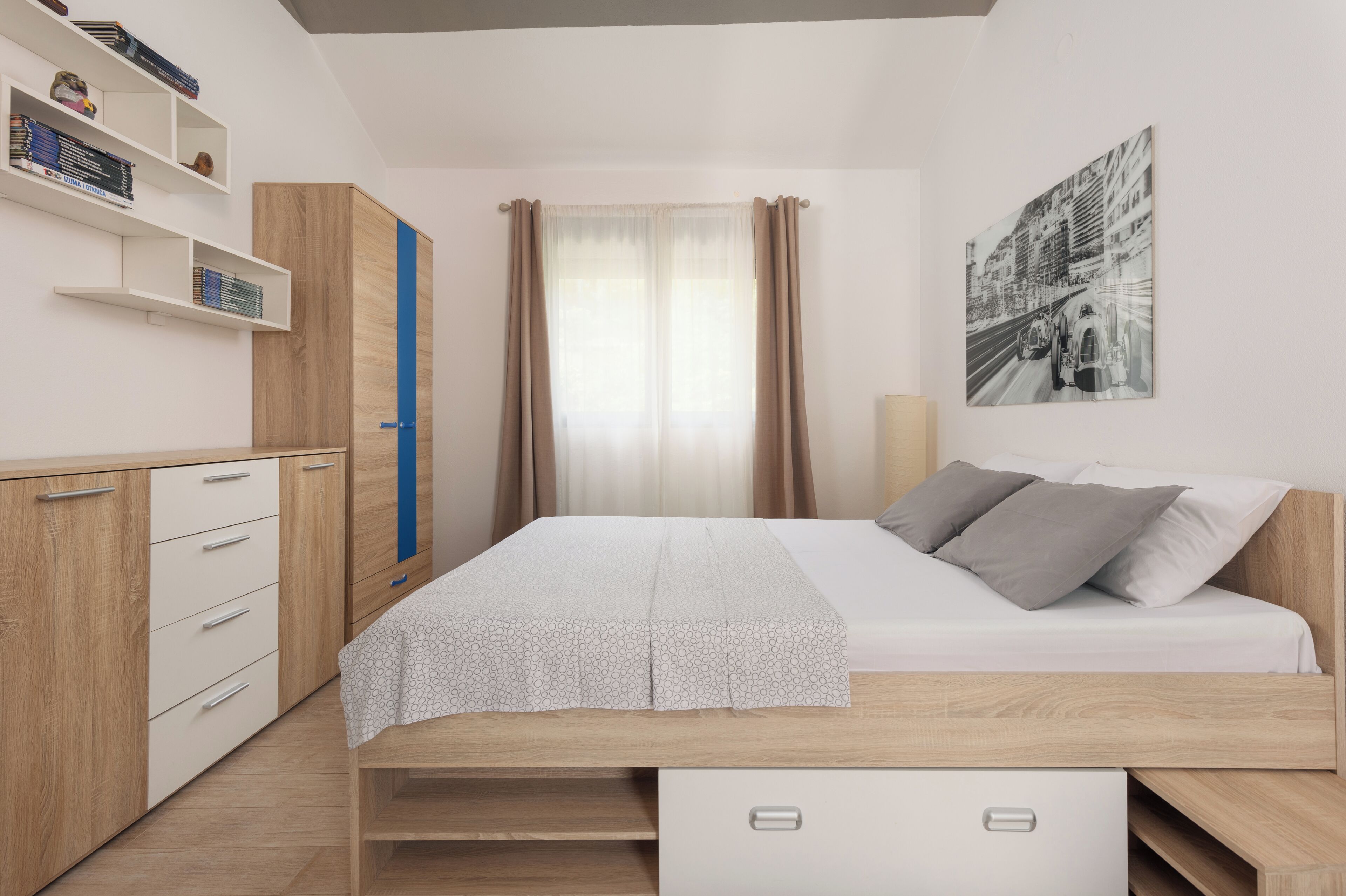A bedroom with a bed, a wardrobe, a bedside table, and a wall-mounted shelf with books.