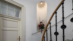 Eine weiße Tür mit Messingklinke, eine Holztreppe mit schwarzen Stäben und ein an der Wand befestigter Spiegel, der einen Blumenstrauß mit roten Blüten reflektiert.