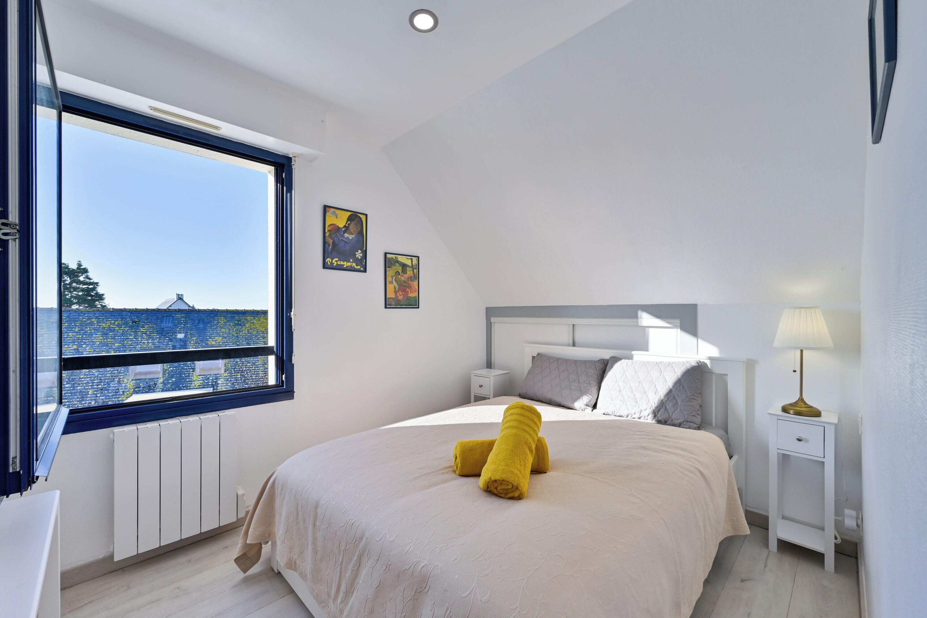 A bedroom with a large window, a bed with a white headboard, a bedside table with a lamp, and a yellow towel on the bed.
