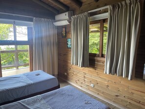 A cabin with two beds, wooden walls, a window with curtains, and an air conditioning unit. - Villa Linda Vista (Malpaís)