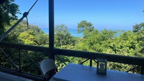 Un balcon avec une table et une chaise, donnant sur une forêt luxuriante et l’océan.