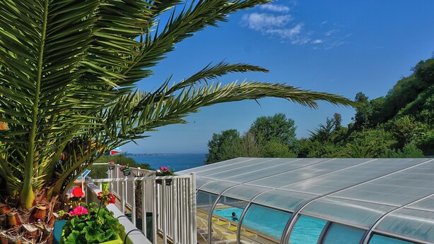 A pool area with a glass enclosure, surrounded by lush greenery and a view of the sea. - Cap - Confort 3 TV – 8p (2) (Plouézec)