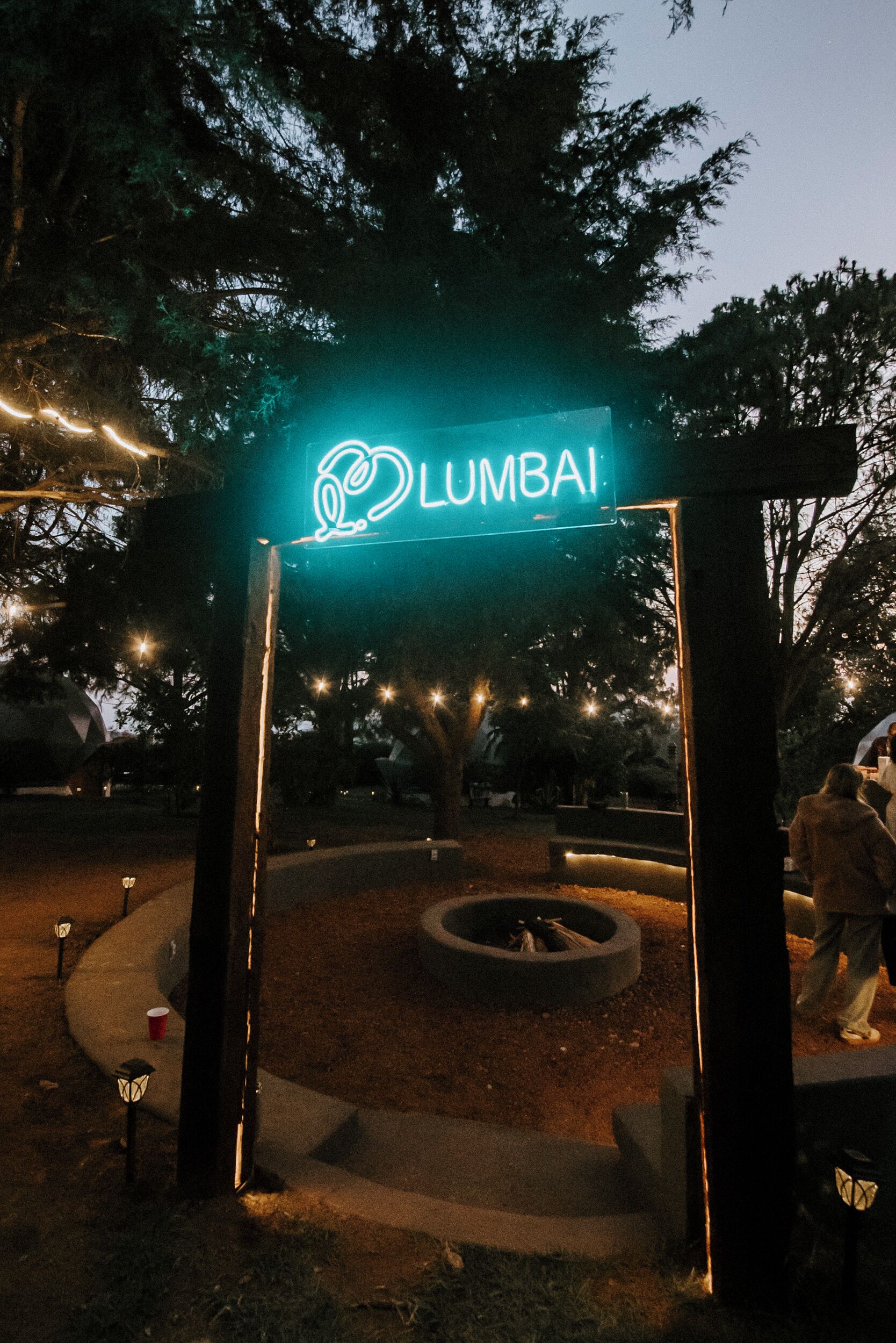 Een nachtzicht van een park met een neonbord waarop 'LUMBAI' staat en een rond zitgedeelte.
