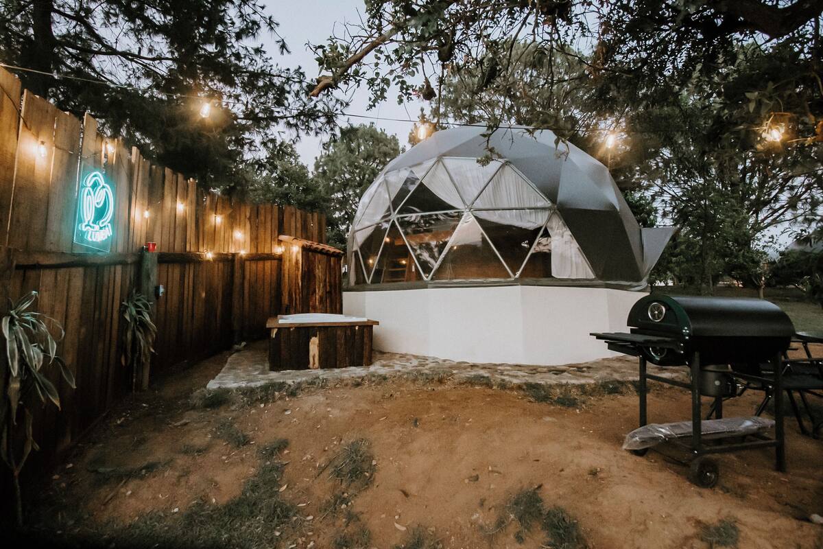 Una estructura de domo geodésico con una parrilla de barbacoa y un letrero de neón en una valla de madera.