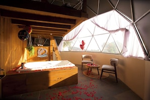 A room with a wooden bathtub, a chair, a small table with a glass, and a window with a view.