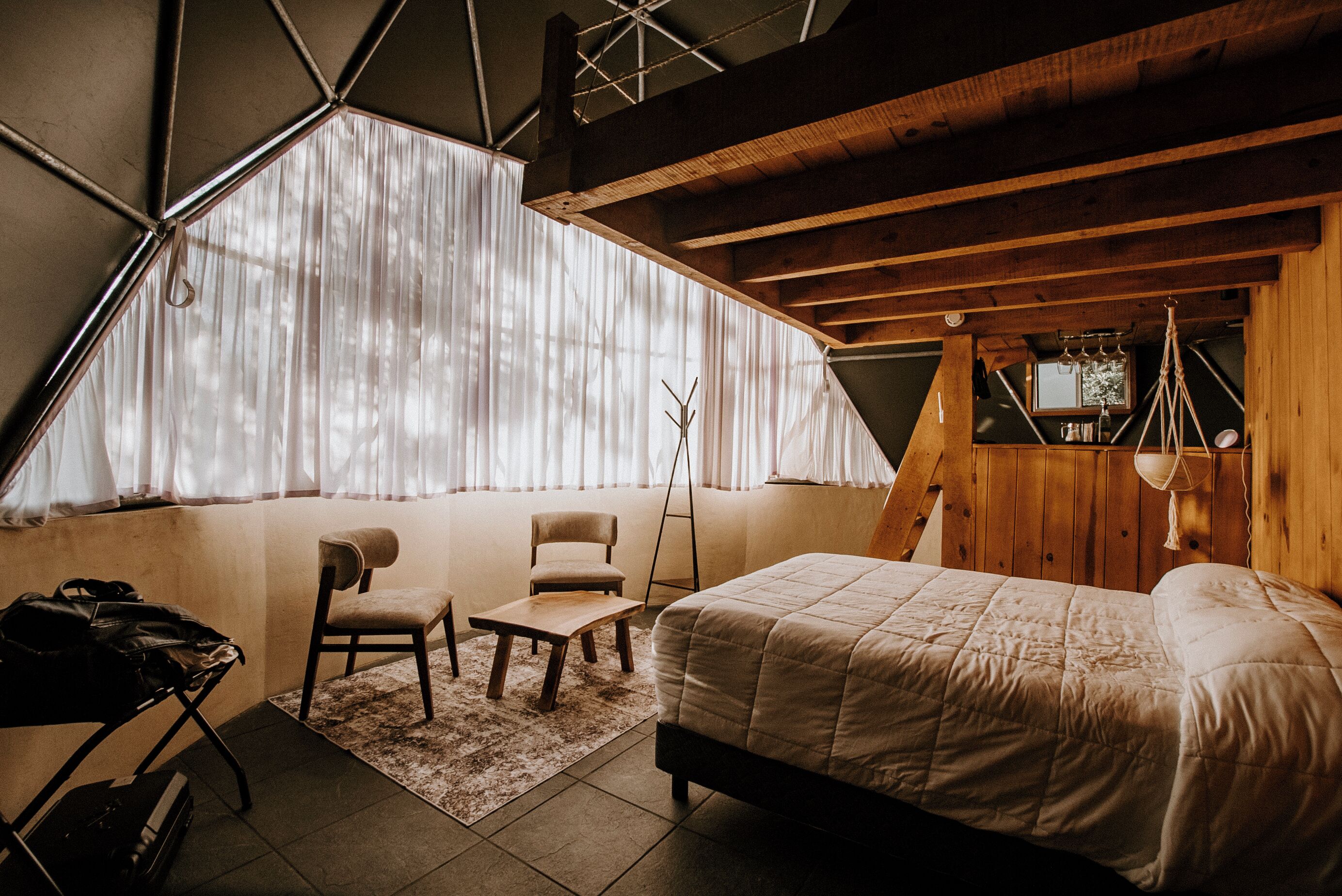 Quarto com uma cama, uma cadeira, uma mesinha e uma janela com cortinas.