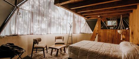 Quarto com uma cama, uma cadeira, uma mesinha e uma janela com cortinas.