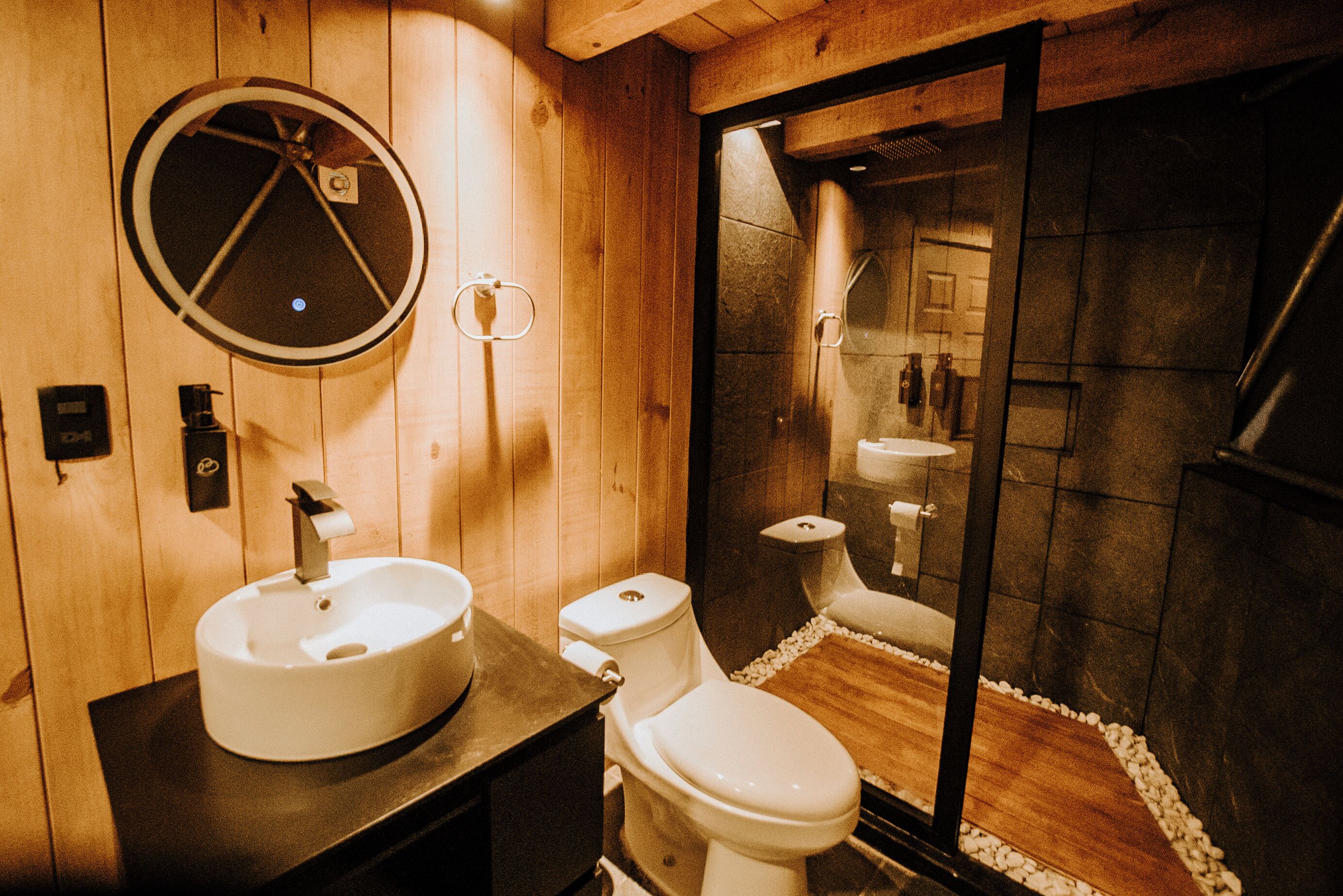 A wooden bathroom with a round mirror, a sink, and a toilet.