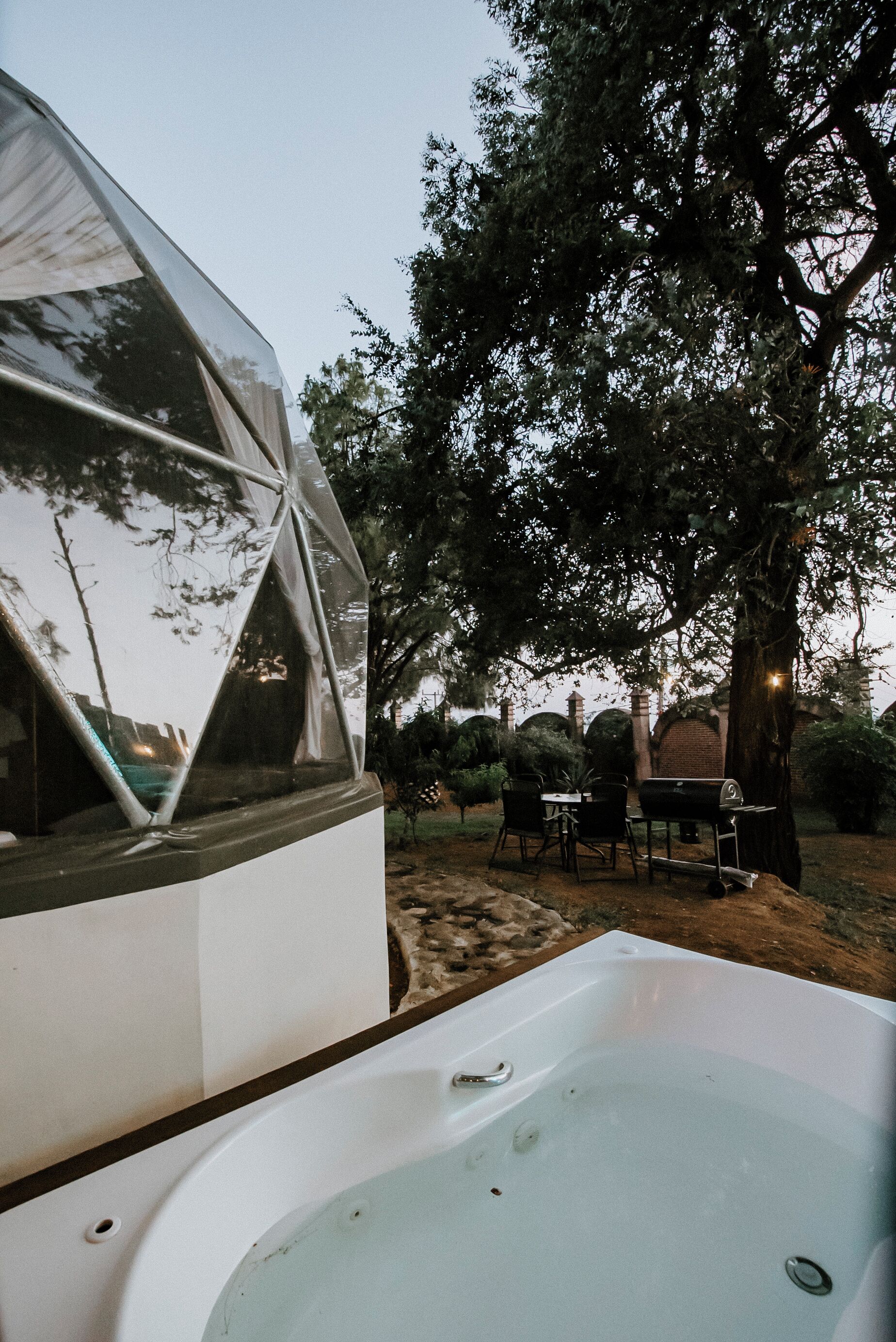 A modern outdoor setting with a hot tub, a dining area, and a large tree.