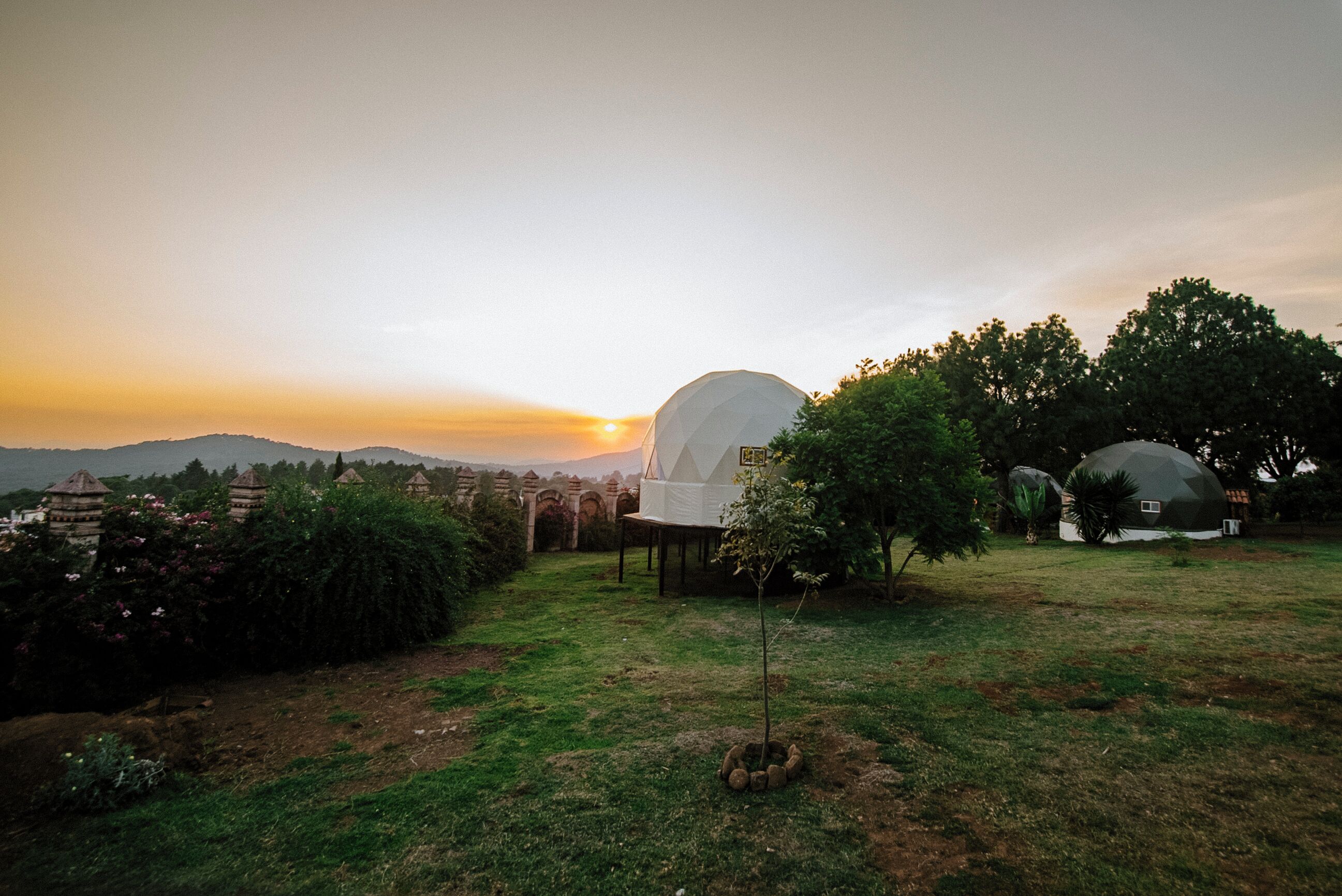 Un bâtiment en forme de dôme offrant une vue dégagée sur le coucher du soleil, entouré de verdure et d’autres constructions similaires.