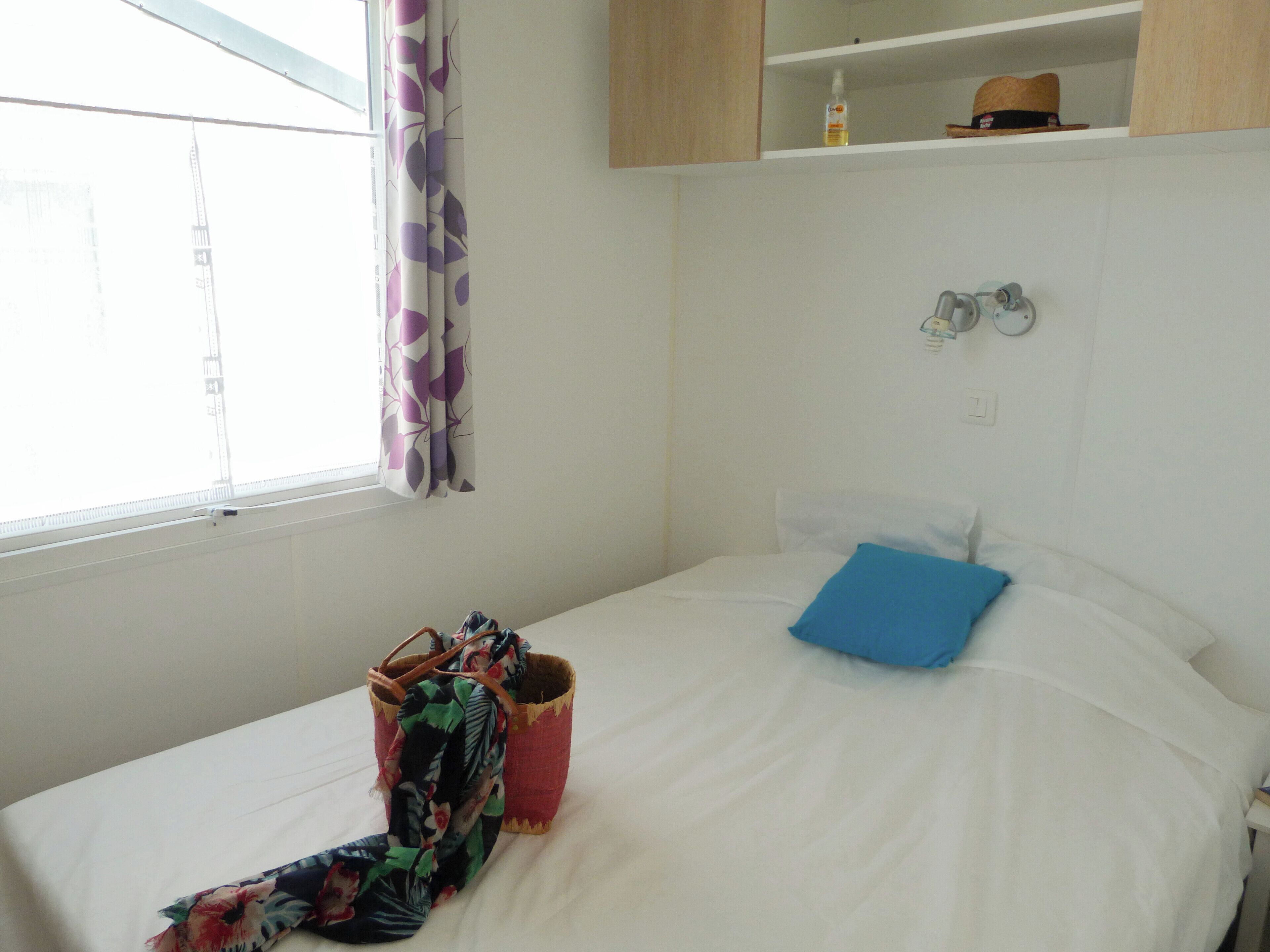 Ein Einzelbett mit blauem Kopfkissen, ein kleiner Nachttisch, eine Wandleuchte und ein Fenster mit Vorhängen.