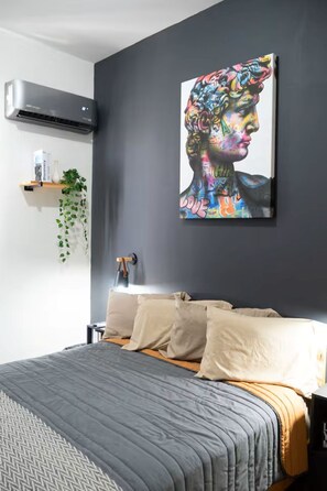 Une chambre à coucher moderne avec un lit gris, des oreillers beiges et un tableau abstrait aux couleurs vives accroché au mur.