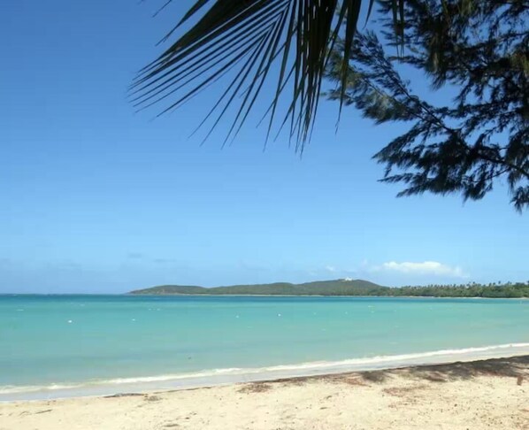 A beach with clear turquoise water, a sandy shore, and a distant mountain range.