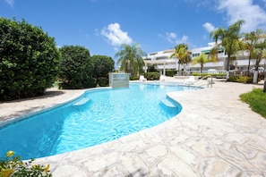 A swimming pool with a stone deck, surrounded by greenery and a building with balconies.