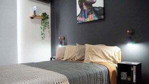 Une chambre moderne avec un lit gris, des oreillers blancs, un plaid à motifs gris et blanc, une petite table de chevet avec une lampe, une étagère murale avec une plante et une œuvre d’art encadrée au-dessus du lit.