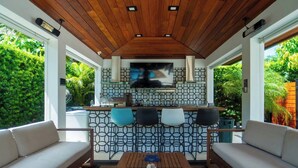 A modern outdoor kitchen with a wooden ceiling, a TV, and a patterned backsplash. There are two sofas and a wooden table in the foreground. - Greenview Villa/Luxury Heated Pool Home Golf View (Miami)