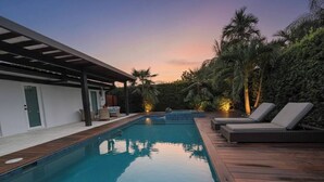 A rectangular swimming pool with a wooden deck and lounge chairs on one side, surrounded by greenery and a covered area with seating. - Greenview Villa/Luxury Heated Pool Home Golf View (Miami)