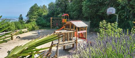 A playground with a basketball hoop and a climbing structure, surrounded by sand and greenery.